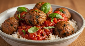 Primer plano de un plato de albóndigas en salsa de tomate sobre arroz blanco, adornado con hojas de albahaca fresca, en un cuenco de cerámica.