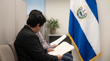 Dos hombres de perfil sentados en sillas de oficina sostienen carpetas con hojas de papel al lado de la bandera salvadoreña.