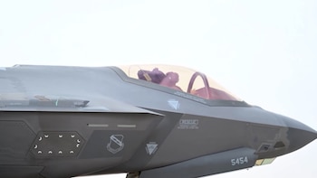 Primer plano de la parte delantera de un avión de combate gris, con un piloto visible en la cabina bajo un dosel tintado. Se ven números '5454' en el fuselaje