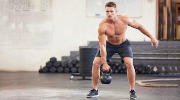 Hombre caucásico, sin camisa, realiza un kettlebell swing con una pesa rusa negra en un gimnasio. Viste pantalones cortos oscuros y zapatillas deportivas.
