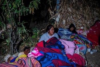 Tres personas, incluyendo un menor, cubiertas con mantas de varios colores, descansan en el suelo al aire libre en la oscuridad junto a árboles y vegetación