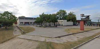 Vista exterior de una escuela de ladrillo claro y techo azul, con un patio pavimentado y árboles frondosos bajo un cielo nublado