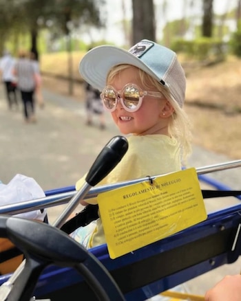 Niña rubia con gorra y gafas de sol redondas, sonríe en un vehículo de pedal azul con un cartel amarillo en italiano. Fondo de árboles y personas