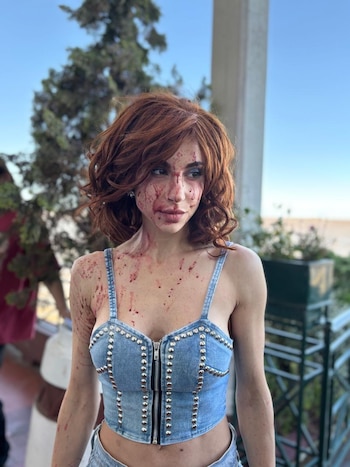 Magui Bravi, una mujer con cabello castaño rojizo y un top de mezclilla con estoperoles, tiene salpicaduras de sangre en la cara y los hombros, mirando hacia la derecha