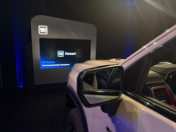 Parte lateral de una camioneta blanca frente a un panel con una pantalla encendida que exhibe el logotipo de General Motors