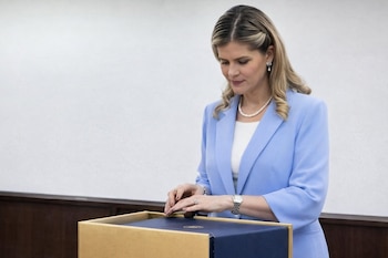 Mujer rubia con blazer azul claro y collar de perlas, inclinada sobre una caja azul oscuro dentro de una estructura dorada, en un entorno formal.