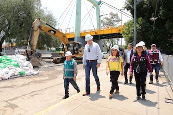 Grupo de personas con cascos y chalecos en una zona de obras. Incluye una excavadora amarilla, pilas de sacos, un puente y árboles en el fondo