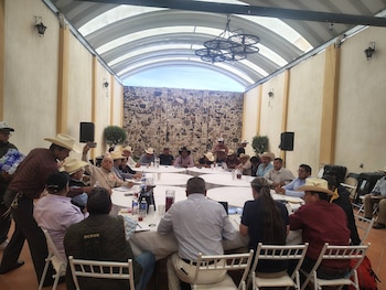 Grupo de hombres, varios con sombreros, sentados en una mesa larga en reunión. Bebidas, cuadernos y laptops en la mesa. Fondo con pared de piedra y techo abovedado translúcido