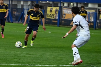 Un jugador de fútbol con uniforme azul y amarillo conduce el balón cerca de un rival vestido de blanco sobre el césped