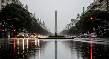 Vista de la Avenida 9 de Julio en Buenos Aires bajo lluvia, con el Obelisco, coches en movimiento, árboles, edificios y charcos que reflejan las luces del tráfico.