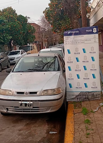 Cartel azul y blanco de la Policía de la Provincia de Buenos Aires, junto a un auto plateado, en una calle con árboles y edificios