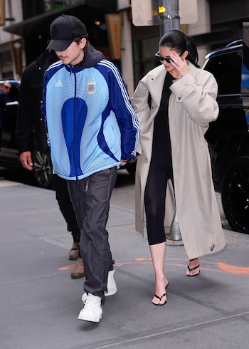 Timothée Chalamet, con gorra negra y chaqueta azul y celeste de Argentina, camina junto a una mujer con gafas de sol, gabardina beige y leggings negros