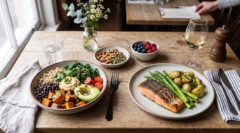 Mesa de madera con un cuenco de granos y verduras, un plato con salmón y espárragos, cuencos con frutos secos y bayas, y una copa de vino.