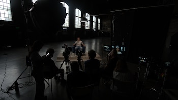 Darren Hayes, vestido con camiseta clara y jeans, sentado en una silla azul en un set de estudio oscuro con grandes ventanales y equipo de cámara