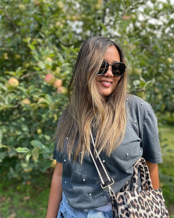 Mujer con cabello largo, gafas de sol negras, camiseta gris con brillantes y bolso estampado de leopardo posa delante de un árbol con frutos