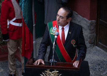 Un hombre con traje oscuro, corbata roja y banda tricolor con escudo, de pie ante un podio con un micrófono. Una persona con uniforme rojo se sitúa a la izquierda
