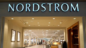 Vista frontal de la entrada de una tienda Nordstrom con su letrero azul brillante, un interior iluminado y productos a la venta