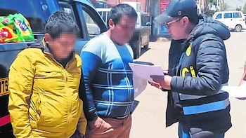 Un agente policial lee un documento frente a dos hombres con el rostro difuminado en una calle junto a un vehículo negro