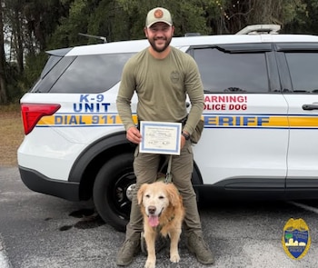 Un oficial de policía sonríe junto a un perro golden retriever frente a una patrulla K-9, sosteniendo un certificado