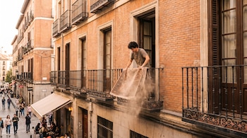 Joven sacudiendo una alfombra desde la ventana de un edificio de ladrillo naranja con balcones sobre una calle con gente y un café.