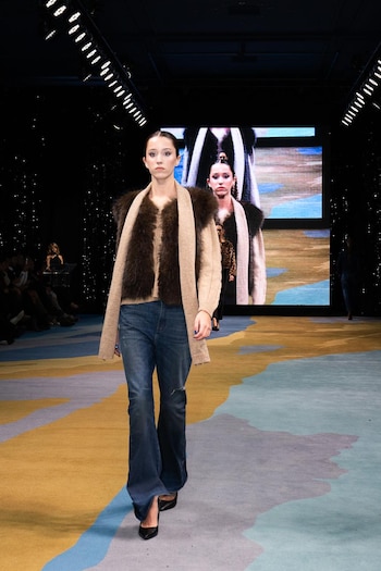 Modelo joven camina por una pasarela con un abrigo de piel sintética marrón, suéter beige, jeans azules acampanados y tacones negros