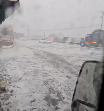 Vista desde el interior de un vehículo de una carretera cubierta de granizo, con coches y un camión en la distancia bajo un cielo nublado y lluvioso