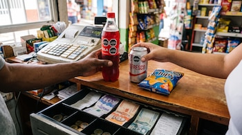 Productos ultraprocesados con sellos de advertencia negros sobre un mostrador de madera junto a una caja registradora con billetes colombianos.