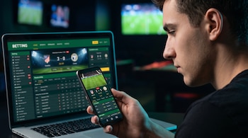 Hombre joven de perfil, mirando un smartphone con app de apuestas de fútbol. Un portátil muestra un sitio de apuestas y pantallas de TV con partidos de fondo.