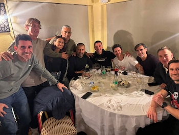 Diez personas posan juntas alrededor de una mesa redonda con mantel blanco, copas, botellas y platos