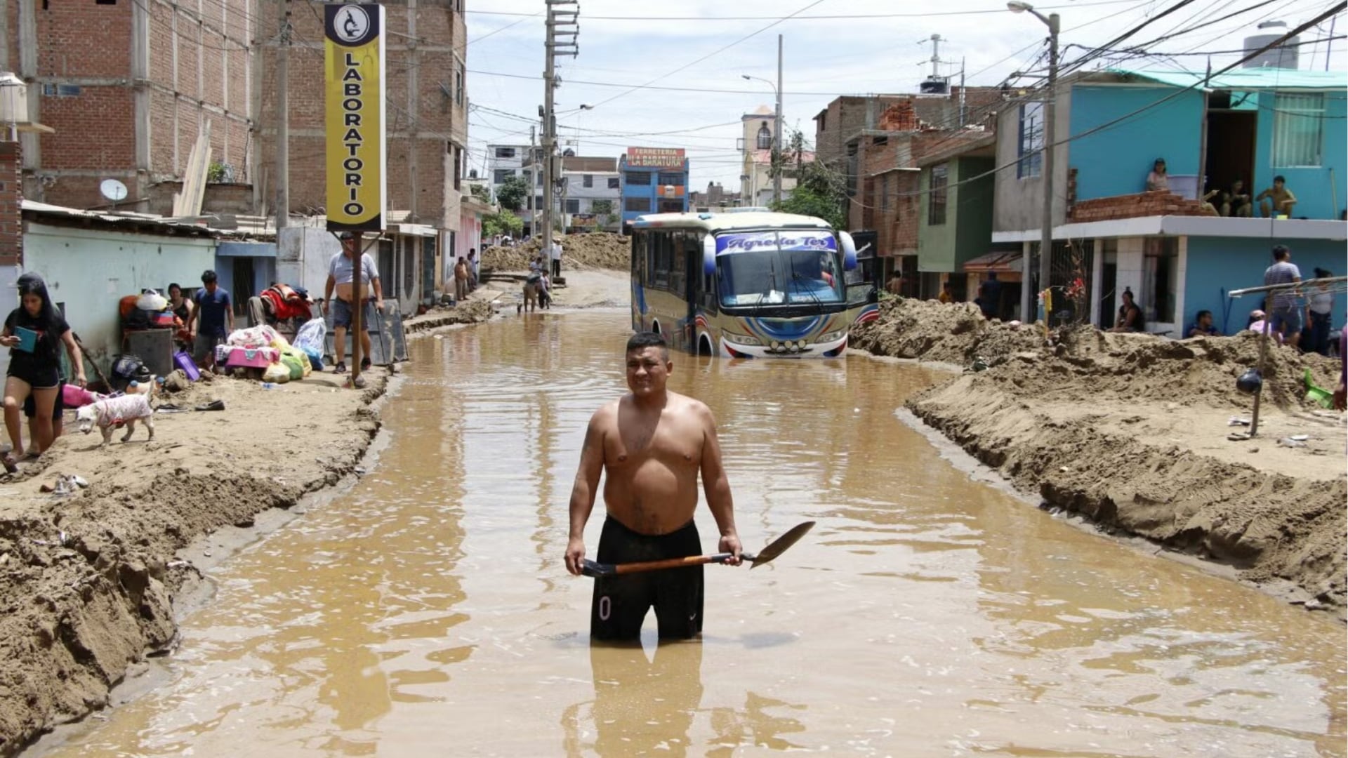 Un hombre con pala avanza por una calle inundada con lodo en Perú, mientras un autobús y habitantes afrontan las consecuencias de las lluvias intensas.