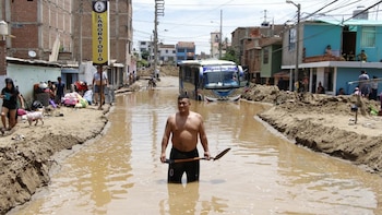 Un hombre sin camiseta sostiene una pala mientras camina en una calle inundada y con lodo, un autobús semihundido y personas se mueven entre edificios