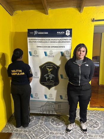 Una mujer de cabello oscuro y chaqueta negra posa para la foto junto a una oficial de policía de espaldas, frente a un cartel oficial