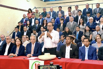 Roberto Sánchez, de pie, habla por un micrófono en un evento político. Está detrás de una mesa roja, con muchas personas sentadas en filas detrás y a su alrededor