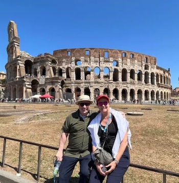 Un hombre con sombrero y una mujer con gorra roja posan sentados ante una valla metálica con el Coliseo al fondo