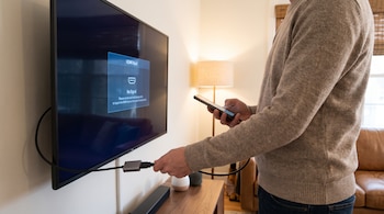 Hombre en suéter gris conecta un cable HDMI con adaptador a un televisor negro que muestra "No Signal", mientras sostiene un teléfono móvil.