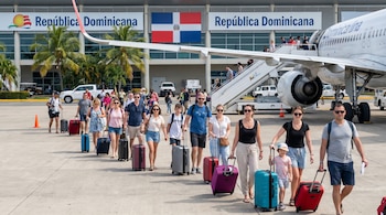 Grupo de turistas con maletas camina por la pista de un aeropuerto soleado en República Dominicana, con un avión y la terminal con la bandera al fondo.