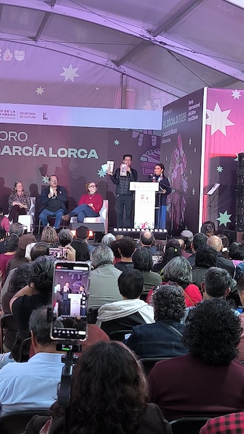 Varias personas presentan un libro sobre un escenario iluminado frente a un público sentado que toma fotos con sus teléfonos