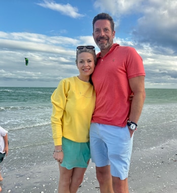Hombre y mujer de pie en una playa arenosa. El hombre usa polo rojo y shorts azules; la mujer, sudadera amarilla y shorts verdes. Mar y cielo nublado al fondo