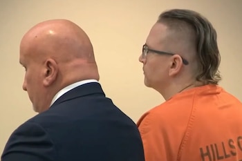 Dos hombres de perfil ante una pared clara, uno de espaldas con traje oscuro y otro con gafas y una vestimenta naranja de recluso