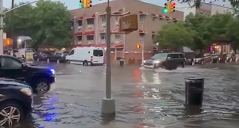 Vista de una intersección urbana anegada con varios coches y una furgoneta blanca avanzando a través del agua. Se ven edificios de ladrillo y semáforos