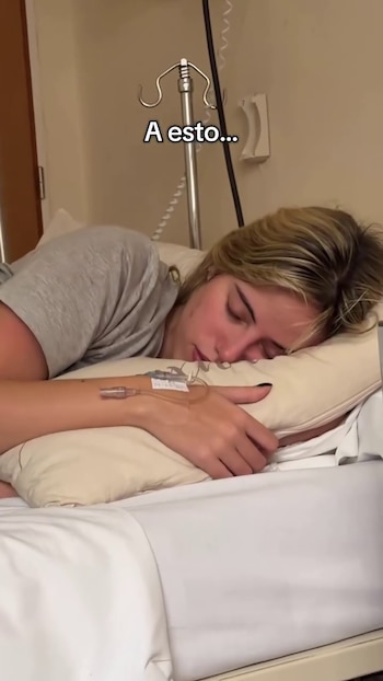 Una mujer rubia yace en una cama de hospital con un soporte de suero cerca. Tiene una vía intravenosa conectada en el brazo izquierdo y descansa sobre una almohada