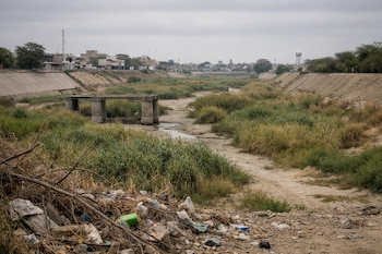 Un río seco con cauce cubierto de vegetación y maleza, un puente de concreto, basura acumulada, terraplenes y edificios de una ciudad bajo cielo nublado.