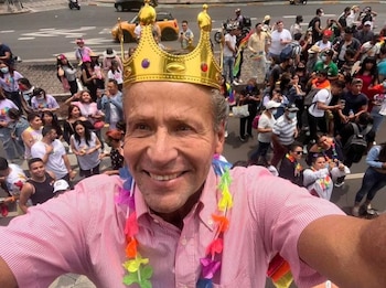 Hombre con camisa rosa a rayas, corona dorada y collar de flores de colores. Detrás, una multitud de personas en la calle