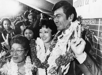 Fotografía en blanco y negro de George R. Ariyoshi sonriendo, rodeado de dos mujeres que también sonríen, todos con leis hawaianos
