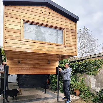 Estructura de madera elevada con ventana y techo oscuro. Dos hombres trabajan en los pilares. Un perro negro y otro claro en el suelo. Fondo con vegetación y cielo nublado