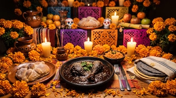 Plato con mole negro y ajonjolí junto a pan de muerto, tortillas, arroz, flores de cempasúchil, velas encendidas y papel picado.