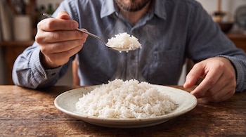Un hombre con camisa a cuadros sostiene una cuchara con arroz blanco caliente sobre un plato hondo en una mesa de madera.