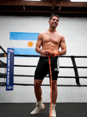 Hombre sin camiseta con vendajes naranjas en las manos y shorts negros, de pie en un ring de boxeo, con bandera argentina y logo Ringside en el fondo