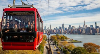 Un teleférico rojo lleno de personas se eleva sobre Roosevelt Island, con el río Este y el horizonte de Manhattan de fondo bajo un cielo azul con nubes.