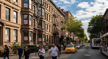 Calle de Nueva York con edificios de ladrillo marrón. Múltiples ventanas muestran carteles de 'Se Alquila'. Personas caminan, taxis amarillos y un autobús transitan por la calle.